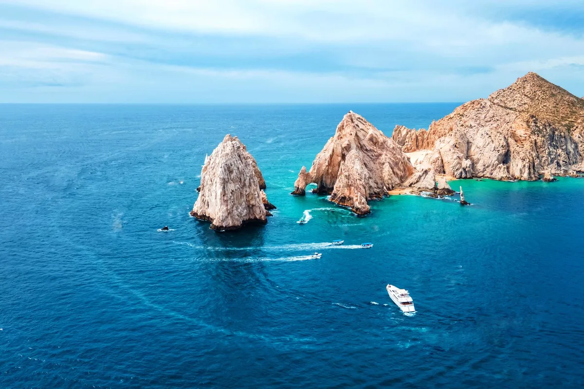 Land's End from the air — the Arch, the sea stacks and a boat's wake in turquoise water