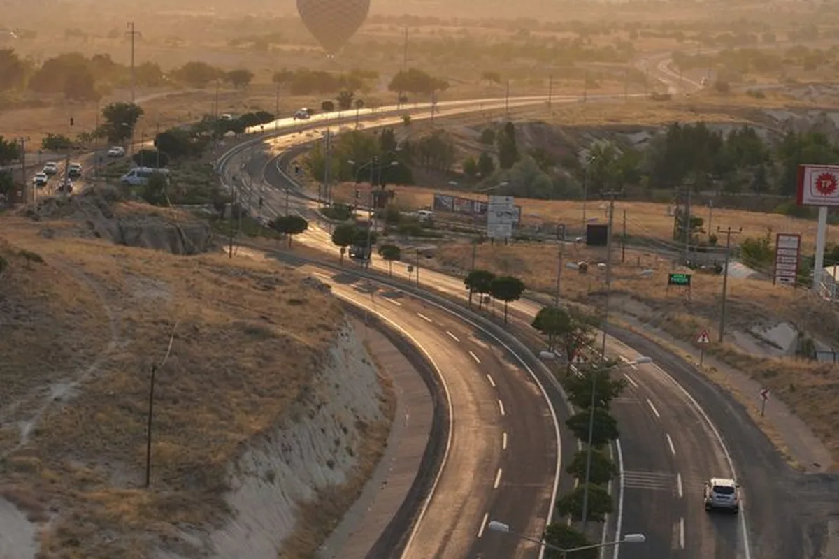 The highway west from Kayseri at dawn, hot air balloons drifting over the valley beside the road