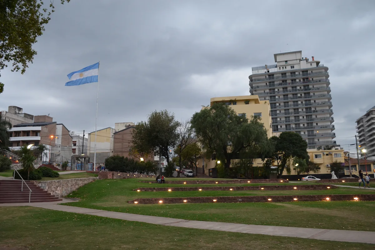 Villa Carlos Paz's central plaza at dusk, the Argentine flag above the gardens