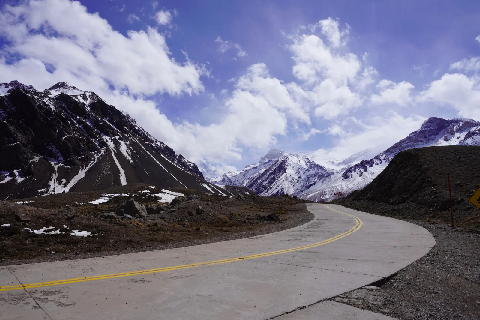 A high alpine road curving past snowfields under big peaks
