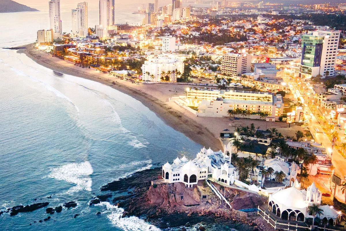 Mazatlán at dusk from above, the malecón curving along the bay and the city lights coming on
