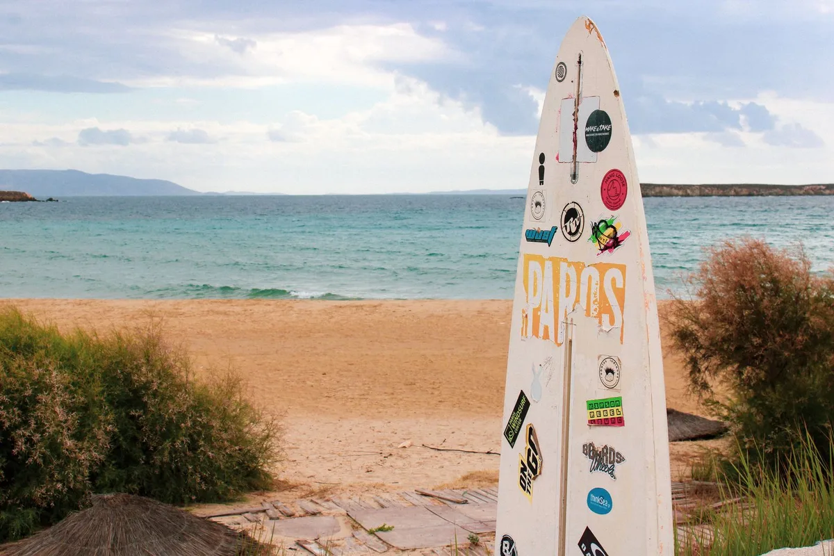 A surfboard covered in stickers standing on a Paros beach, with the Aegean behind it