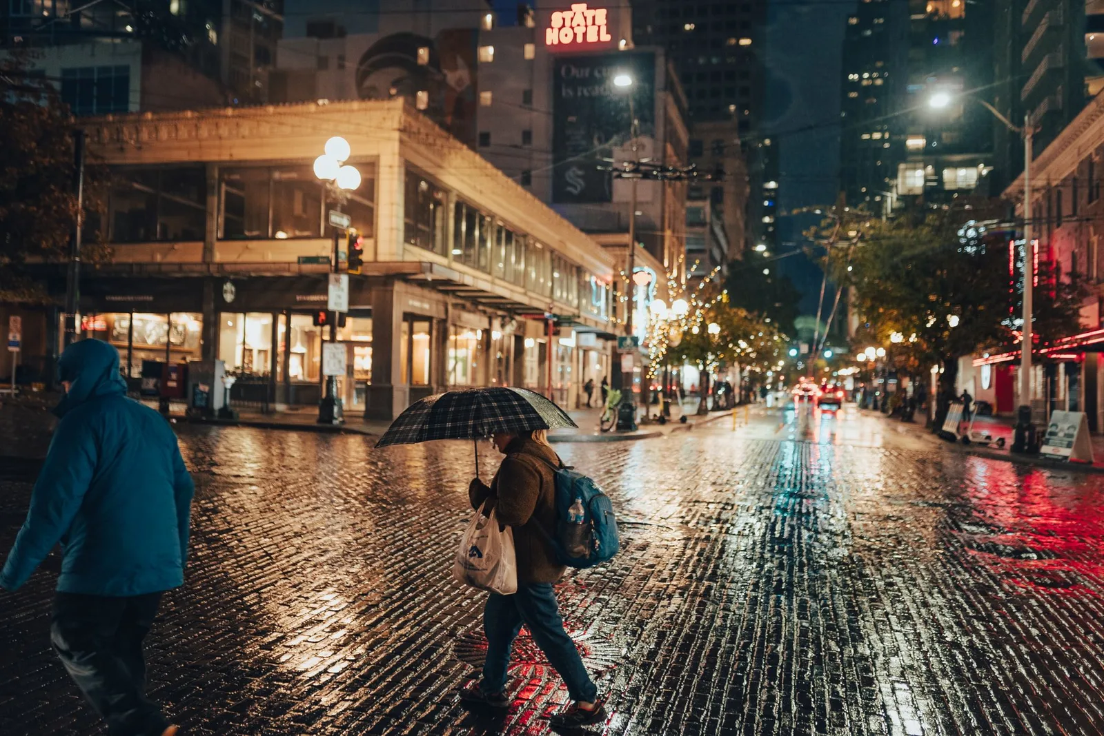 Pike Place in the rain at night, cobblestones shining, a shopper under an umbrella