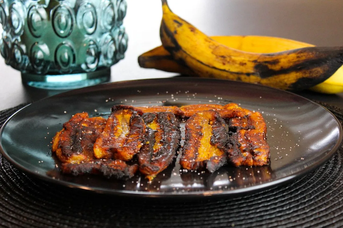 Caramelized fried plantains on a dark plate, a ripe plantain behind