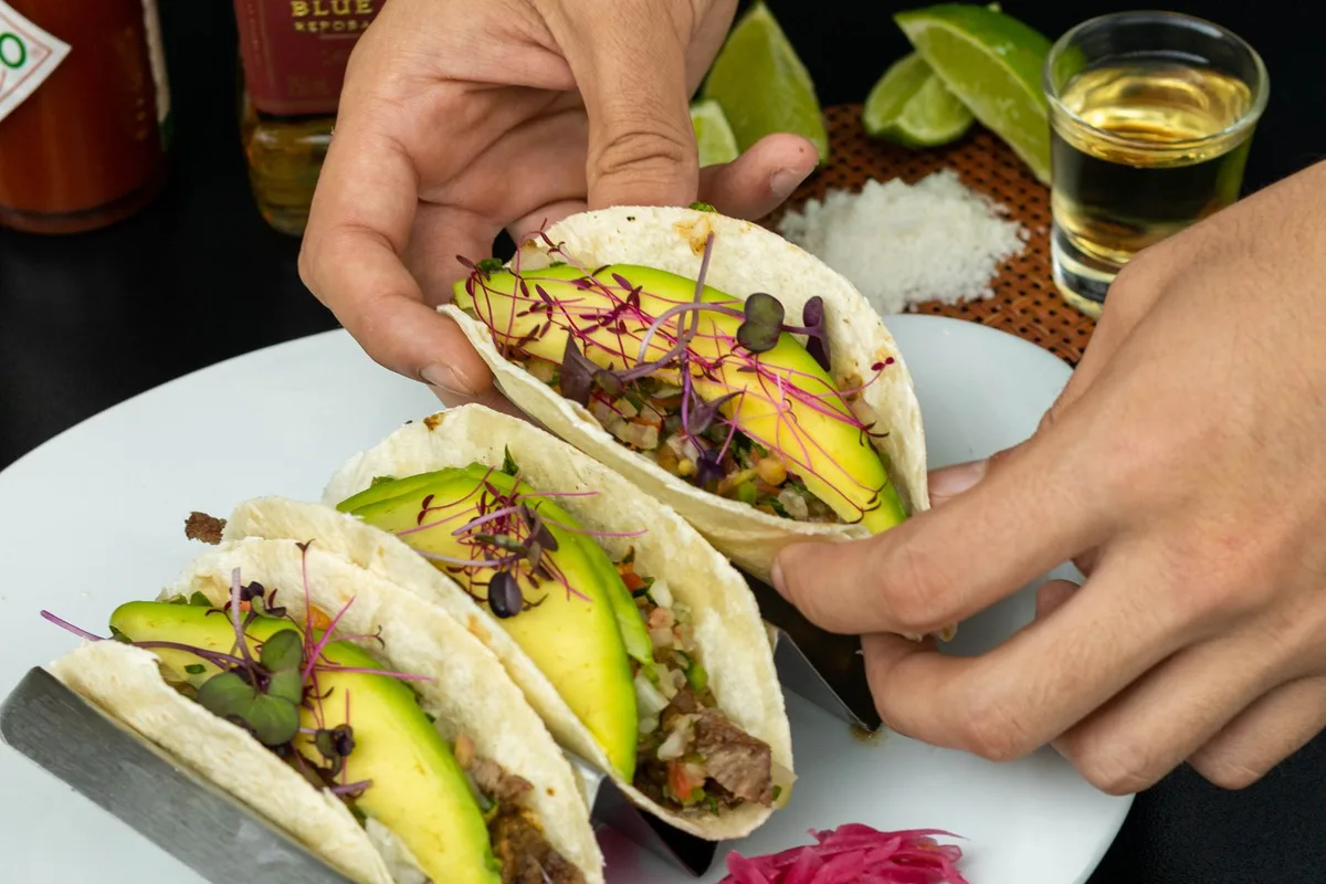Hands lifting tacos over a plate with pickled onion, avocado and lime