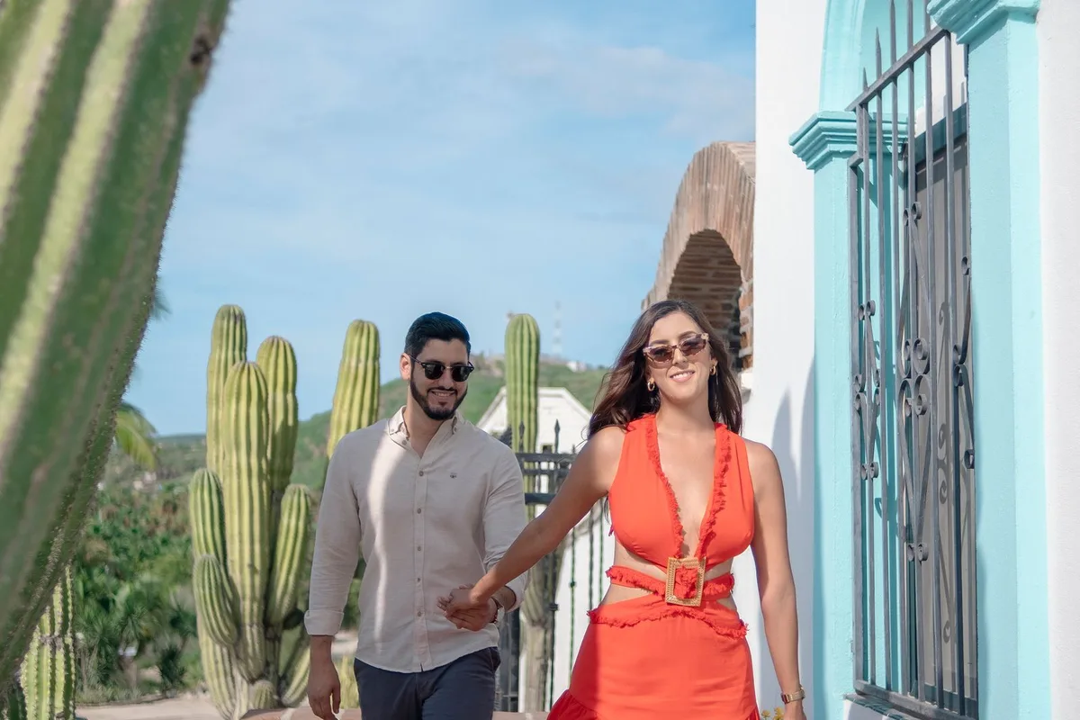 A couple walking past a turquoise colonial facade and tall cactus in Todos Santos